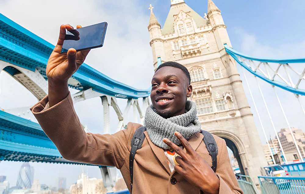 Un bărbat zâmbind care face un selfie cu smartphone-ul său pe fundalul Tower Bridge din Londra.