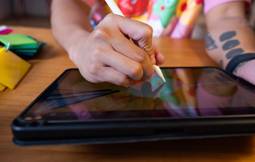 O mână desenează pe o tabletă grafică cu un stylus; piese colorate se află lângă ea