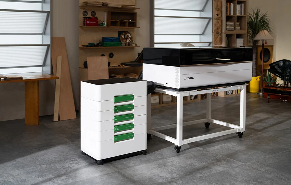 Gravură laser xTool pe un cărucior alb lângă un absorbitor de fum alb cu elemente verzi în atelier