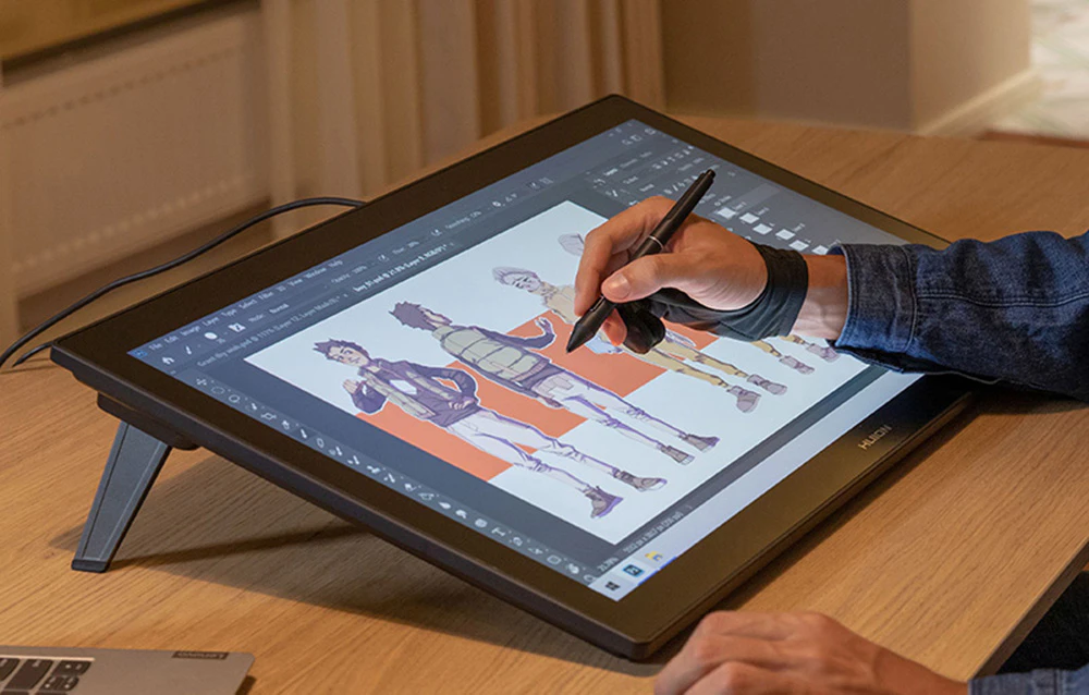 Tabletă Huion pe birou cu suport, persoană desenând caractere în programul grafic, laptop în fundal