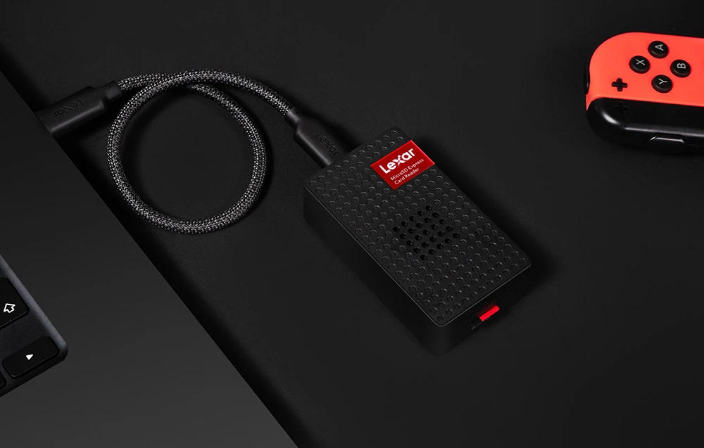 Cititor de carduri Lexar conectat printr-un cablu USB-C la un laptop, așezat pe un birou negru lângă un controler de consolă