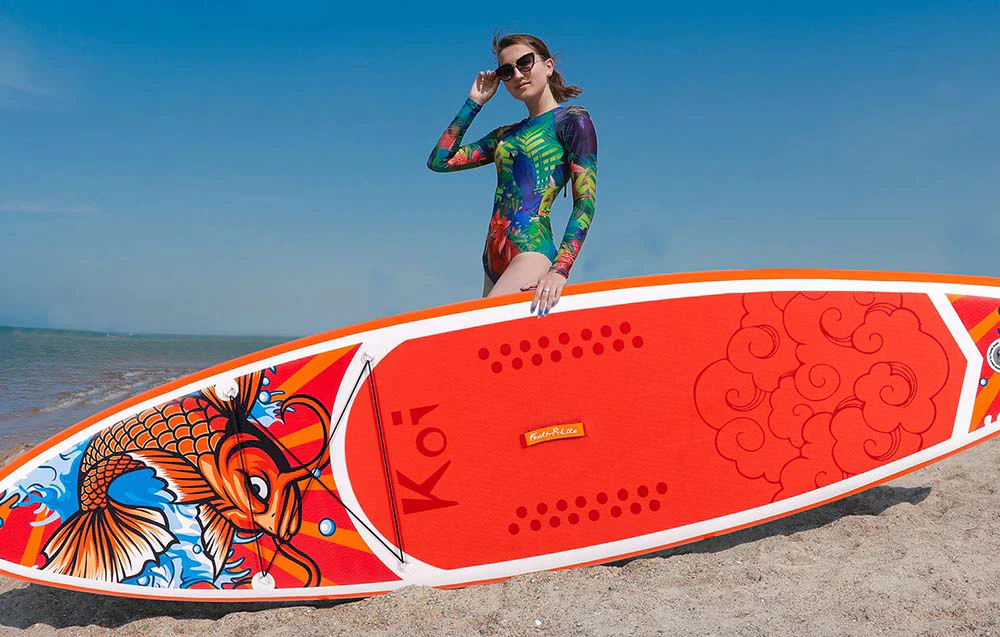 O femeie în costum de baie colorat și ochelari de soare ține cu mâna o placă de surf roșie, în timp ce stă întinsă pe plajă