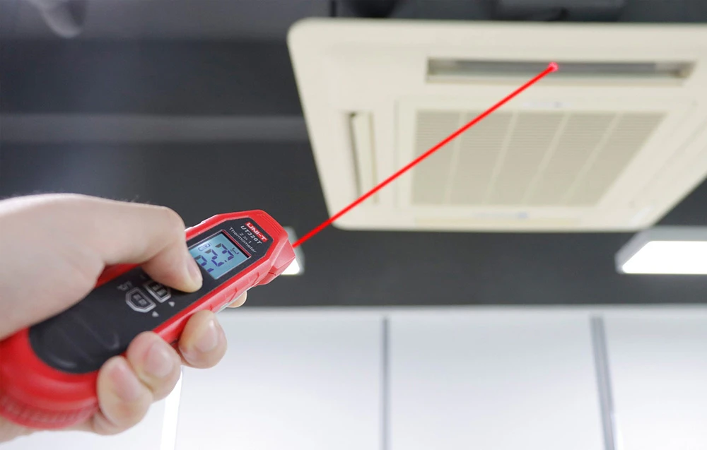 Mână ținând un termometru electronic cu un indicator laser activat și îndreptat către un aparat de aer condiționat de tavan