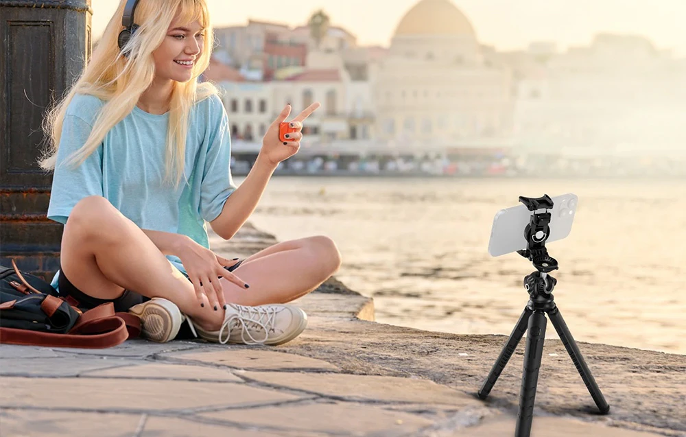 O femeie așezată lângă apă, folosind un trepied pentru a filma cu smartphone-ul pe fundalul unei panorame urbane.