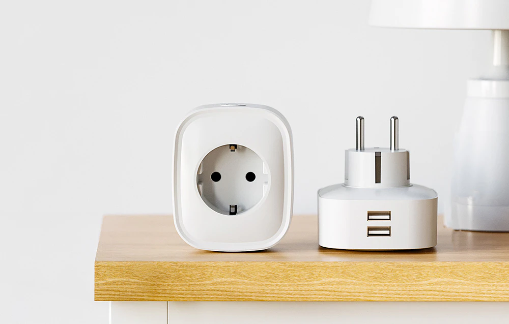 Două adaptoare albe pe blat – unul cu o priză electrică, celălalt cu două porturi USB