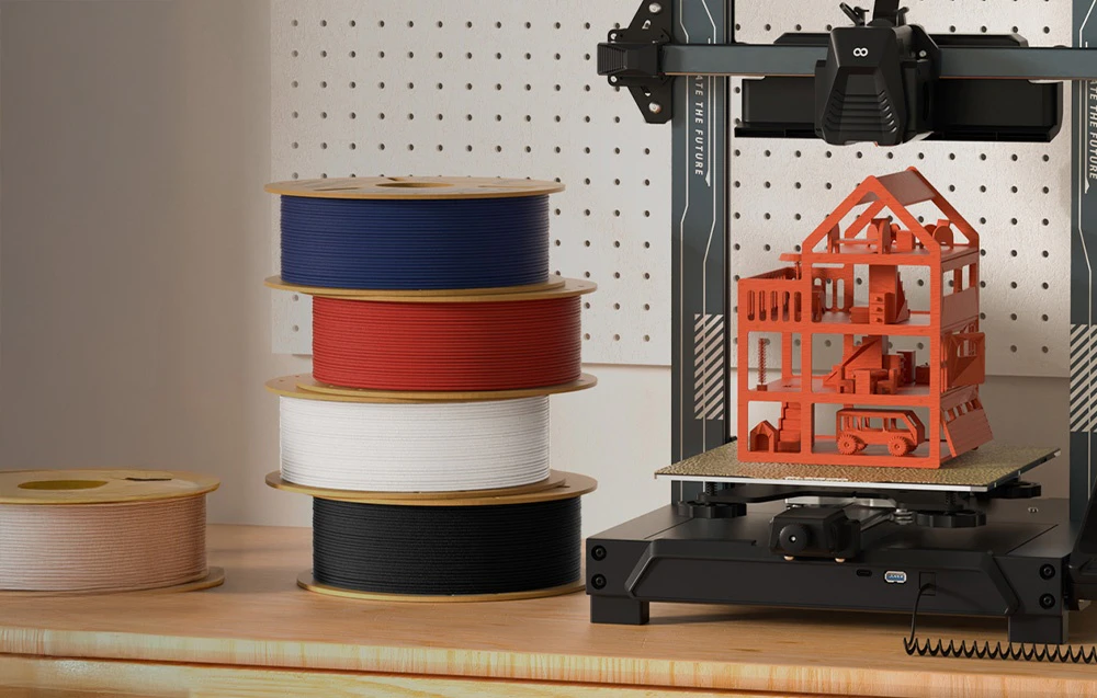 Imprimantă 3D cu o căsuță pentru păpuși de culoare roșie pe platformă, alături de un teanc de mosoare colorate de filament