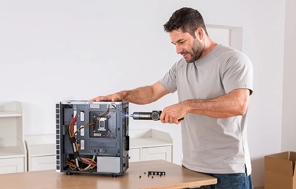 Un bărbat în tricou gri și blugi repară un computer cu o șurubelniță electrică.