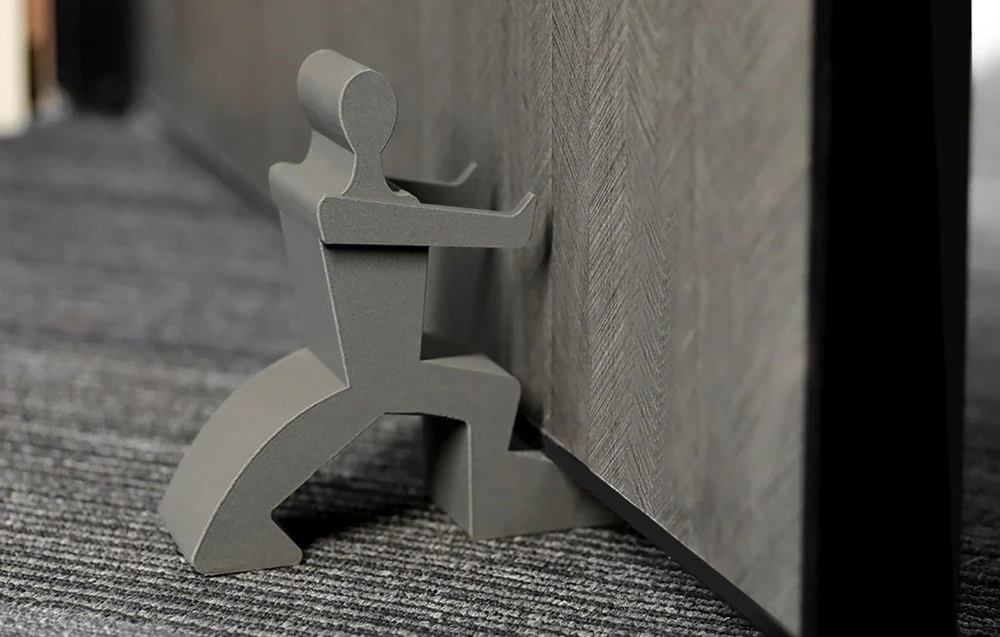 O figurină gri imprimată 3D servește drept pârghie pentru a ține deschisă o ușă așezată pe covor