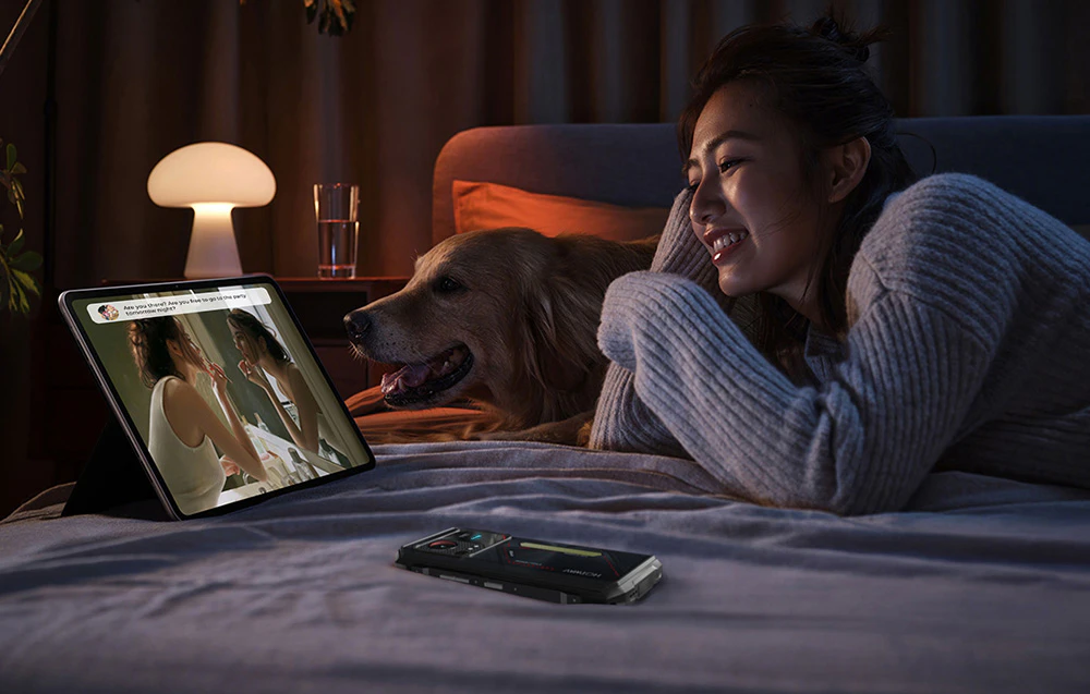 O femeie întinsă pe pat cu câinele ei, urmărind un videoclip pe o tabletă; lângă ea, pe plapumă, se află un smartphone HOTVWAV.