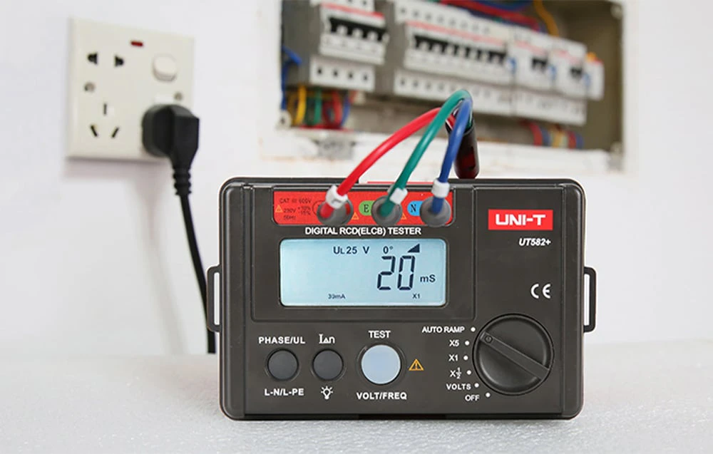 Tester digital RCD UNI-T pe o masă, cu cabluri conectate. O priză de alimentare și un panou de distribuție în fundal.