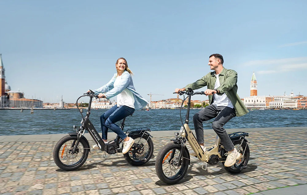 O femeie și un bărbat mergând cu biciclete electrice de-a lungul unei promenade de pe malul apei