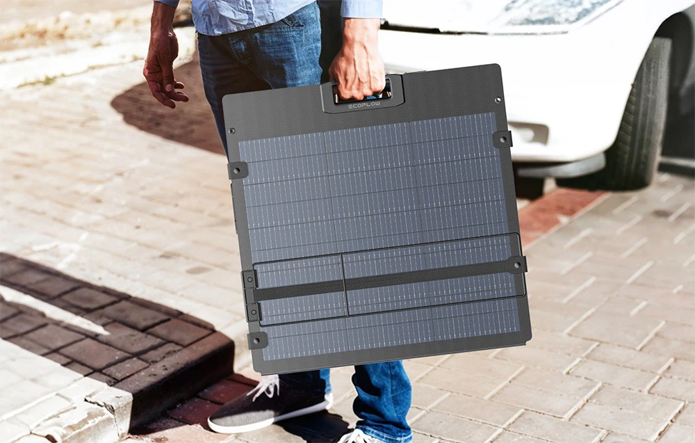 Un bărbat transportă un panou solar pliat cu mânerul în mână, lângă o mașină albă