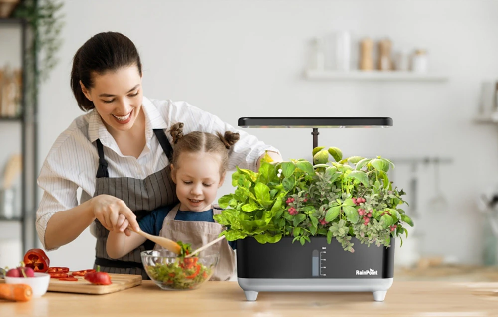 O femeie și un copil pregătesc o salată într-un bol lângă o grădină hidroponică de acasă cu plante aromatice.