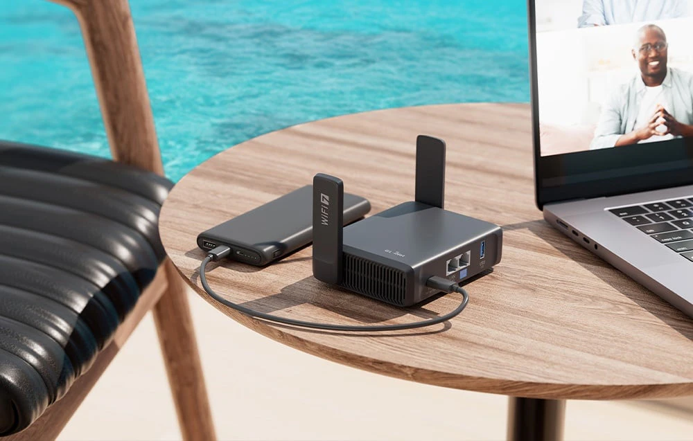 Un router negru conectat la o baterie externă se află pe masa de lângă laptop; în fundal se vede o piscină