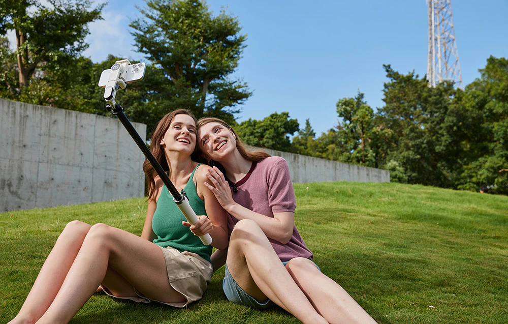 Două femei stau pe iarba dintr-un parc și își fac un selfie cu un gimbal ținut cu brațul întins.