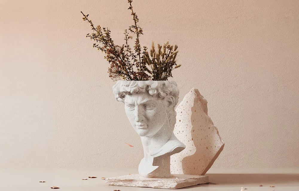 O sculptură albă reprezentând un cap, folosită ca vază pentru plante uscate, așezată pe un fundal deschis, lângă o bucată de stâncă.