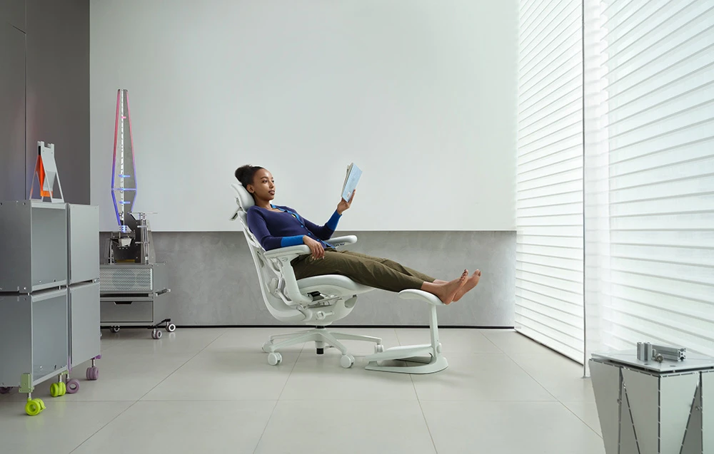 O femeie îmbrăcată într-un pulover violet citește o carte într-un scaun ergonomic alb cu suport pentru picioare, într-o cameră luminoasă