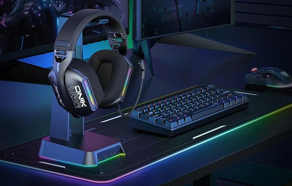 Căști de gaming agățate pe un suport, lângă o tastatură, un mouse și două monitoare