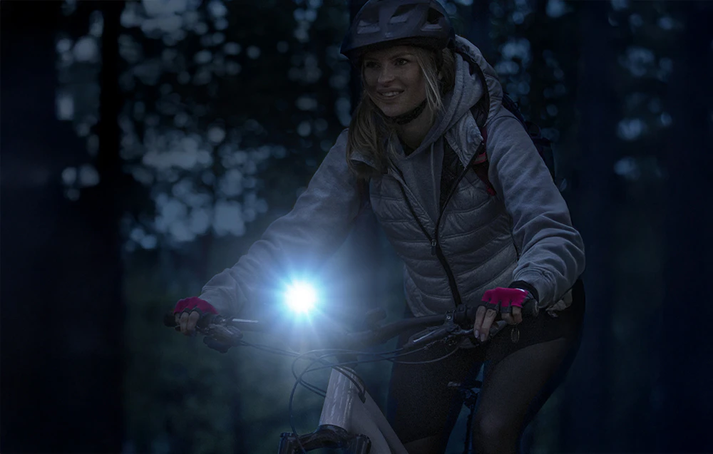 O femeie purtând cască, mergând cu bicicleta noaptea prin pădure, luminând drumul cu o lanternă montată pe ghidon.
