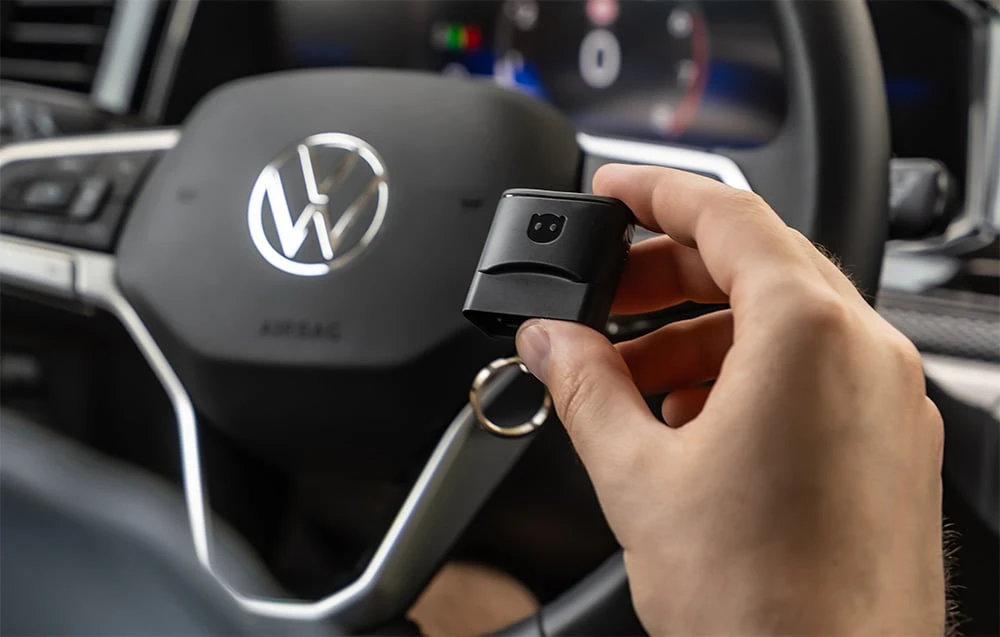 O mână care ține adaptorul Carly în fața unui volan Volkswagen cu logo-ul VW vizibil