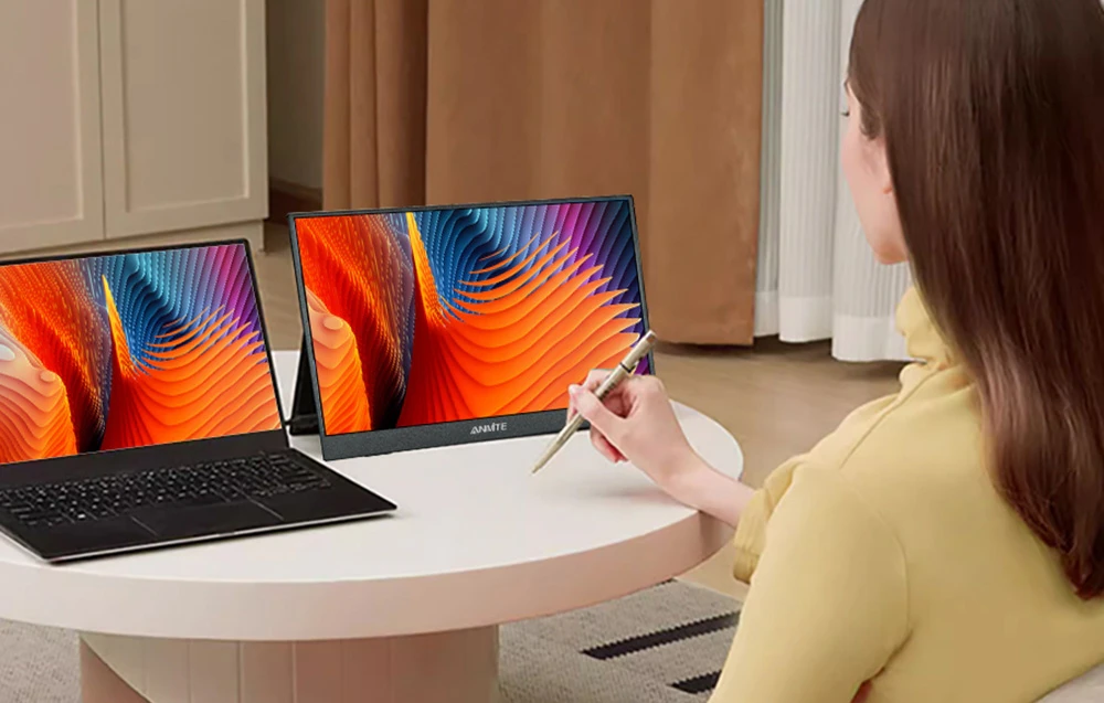 O femeie așezată la o masă rotundă folosește un stylus, având lângă ea un ecran ANMITE suplimentar, amplasat lângă un laptop deschis.