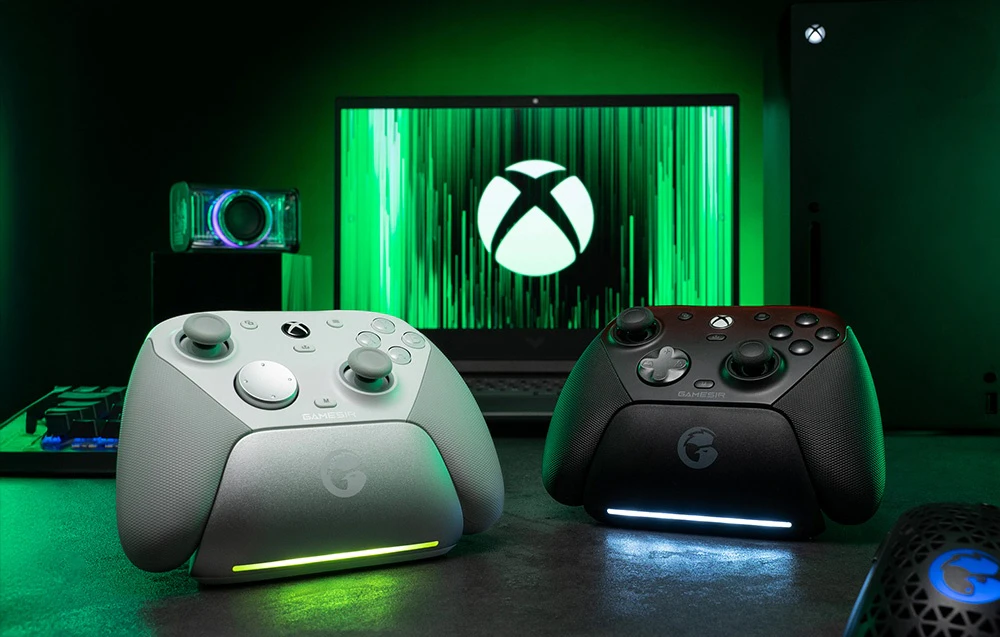Două controlere GameSir pe o stație de andocare în fața unui monitor cu logo-ul Xbox, unul alb, celălalt negru.