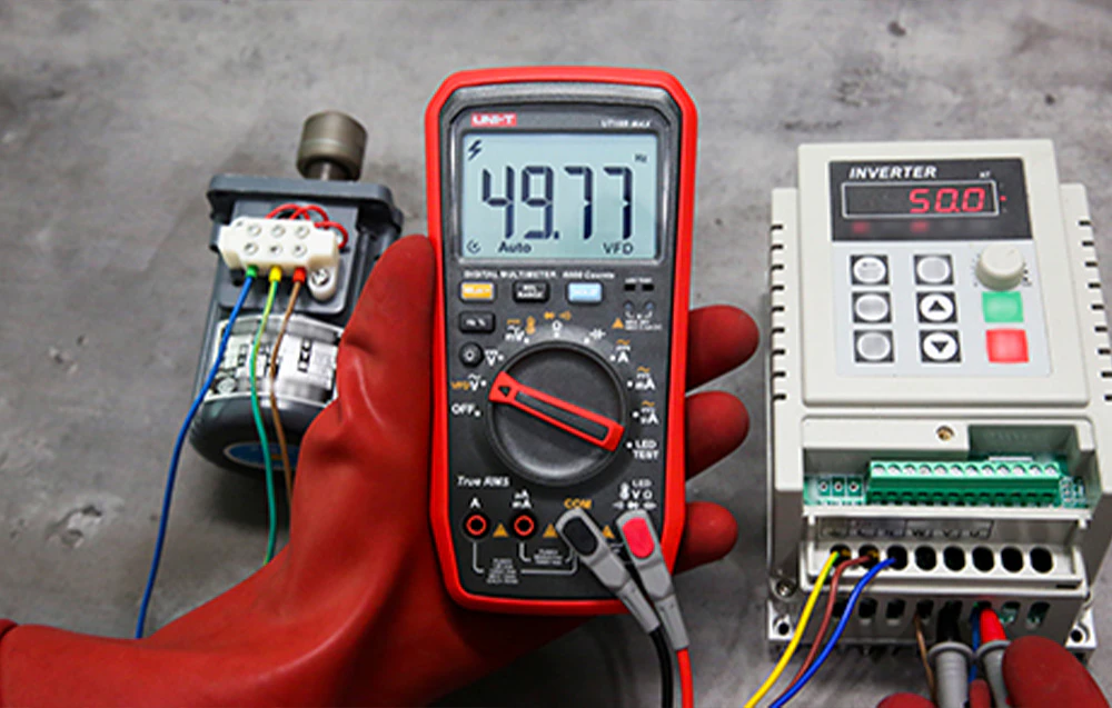 O mână într-o mănușă roșie ținând un multimetru UNI-T care afișează 49,77 Hz, poziționat lângă un motor și un invertor.