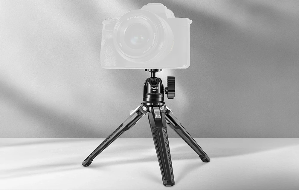 Un mini trepied stă pe o masă albă, susținând o cameră foto mirrorless de dimensiuni normale, cu obiectiv.