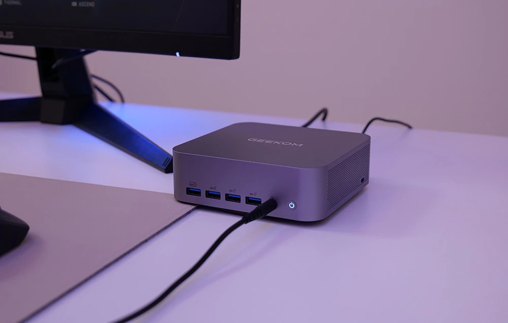Un MINI PC Geekom gri, cu un cablu conectat la el, este așezat pe un birou lângă un monitor într-o cameră cu iluminare violetă