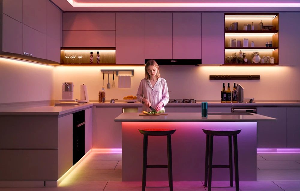 O femeie în pijamale taie legume la insula din bucătărie; în spatele ei, mobilier luminos iluminat de benzi LED colorate