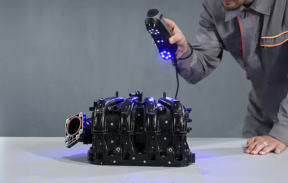 O persoană scanează partea neagră a unui motor cu un scaner 3D portabil cu lumină albastră.