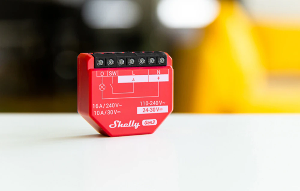 Modulul roșu Shelly 1PM Gen3 stă pe o suprafață albă, cu marcaje tehnice și conectori vizibili din față