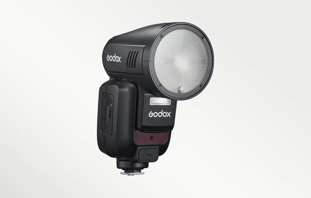 Un bliț Godox V100 negru cu cap rotund, prezentat din față pe un fundal de culoare deschisă