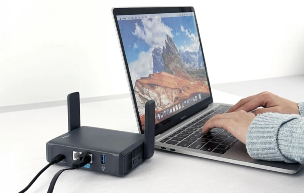 O persoană tastează pe tastatura unui laptop; un router negru se află pe masa de lângă acesta
