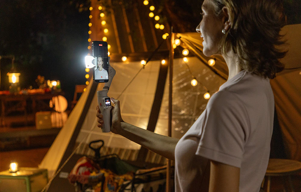 O femeie se înregistrează cu un smartphone montat pe un cardan cu o lumină LED în fața unui cort cu ghirlande de lumină.