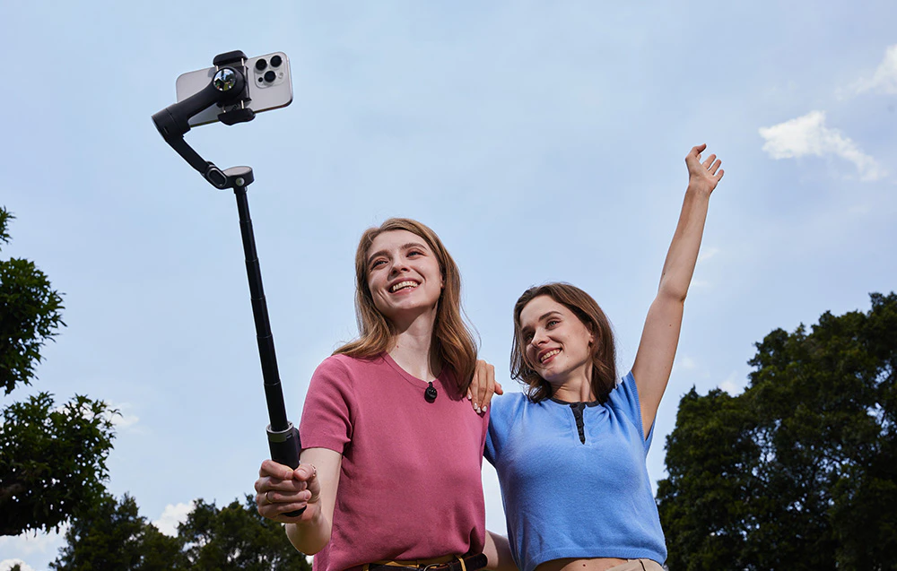 Două femei își fac un selfie împreună în aer liber, folosind un gimbal cu braț extensibil.