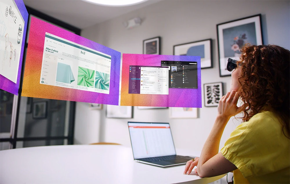 O femeie cu ochelari XR lucrează la un birou cu un laptop, uitându-se la ecrane virtuale în aer