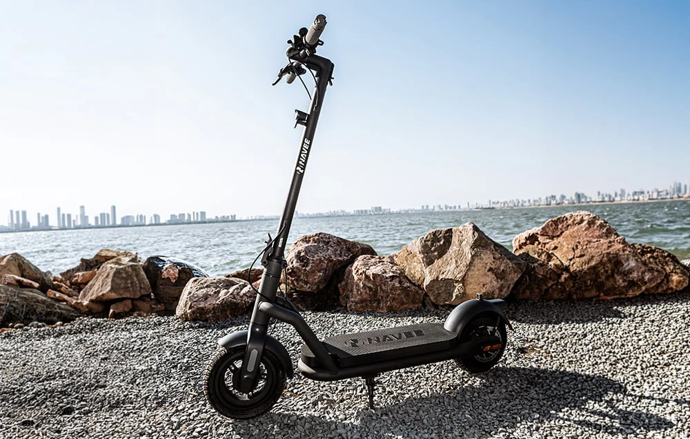 Un scuter electric Navee negru parcat pe malul stâncos, cu apa și orizontul orașului în fundal.