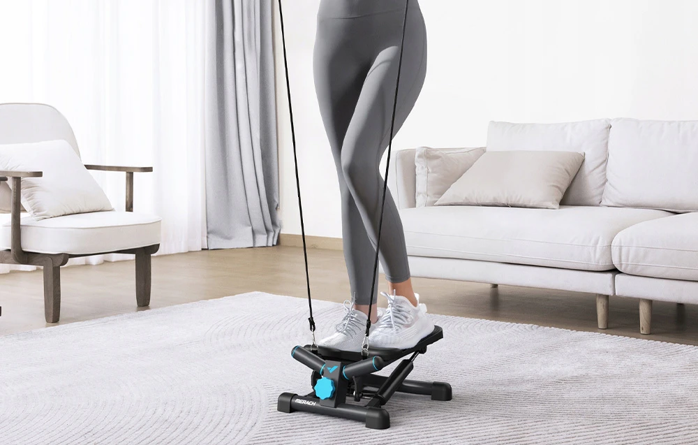 O femeie face exerciții fizice într-un living pe un stepper MERACH negru cu accente albastre, ținând în mâini benzi de exerciții