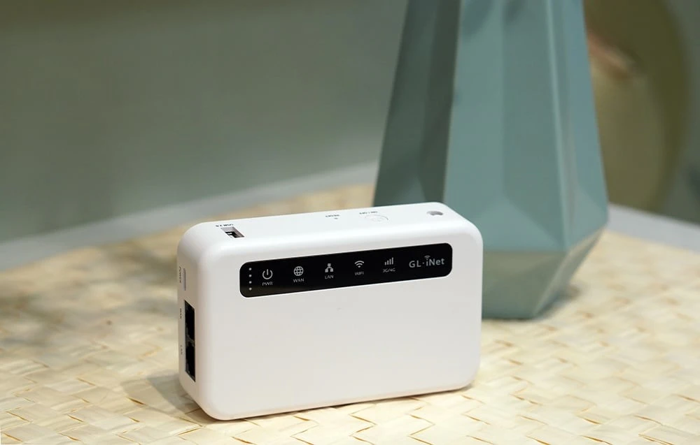 Un router alb așezat în poziție verticală pe un suport împletit, lângă o vază cu flori și decorațiuni din sticlă.