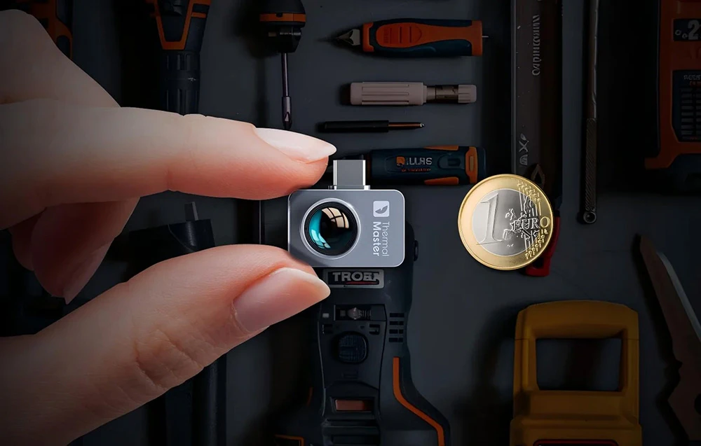 Degetele ținând mini-camera termică Thermal Master lângă o monedă de 1 euro, pe fundalul unor unelte de atelier