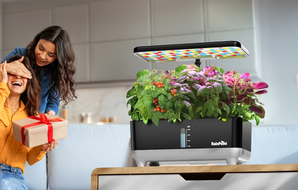 Pe masa de lângă două femei și un cadou se află un sistem hidroponic cu un panou LED colorat aprins