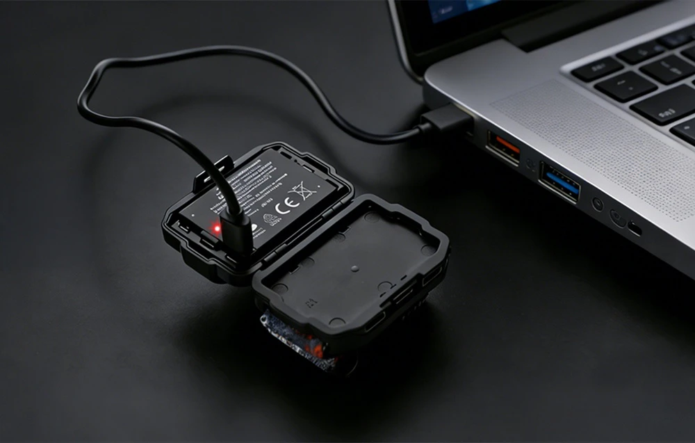 Modulul de baterie deschis al lanternei frontale este conectat printr-un cablu USB negru la portul lateral al unui laptop pentru încărcare.