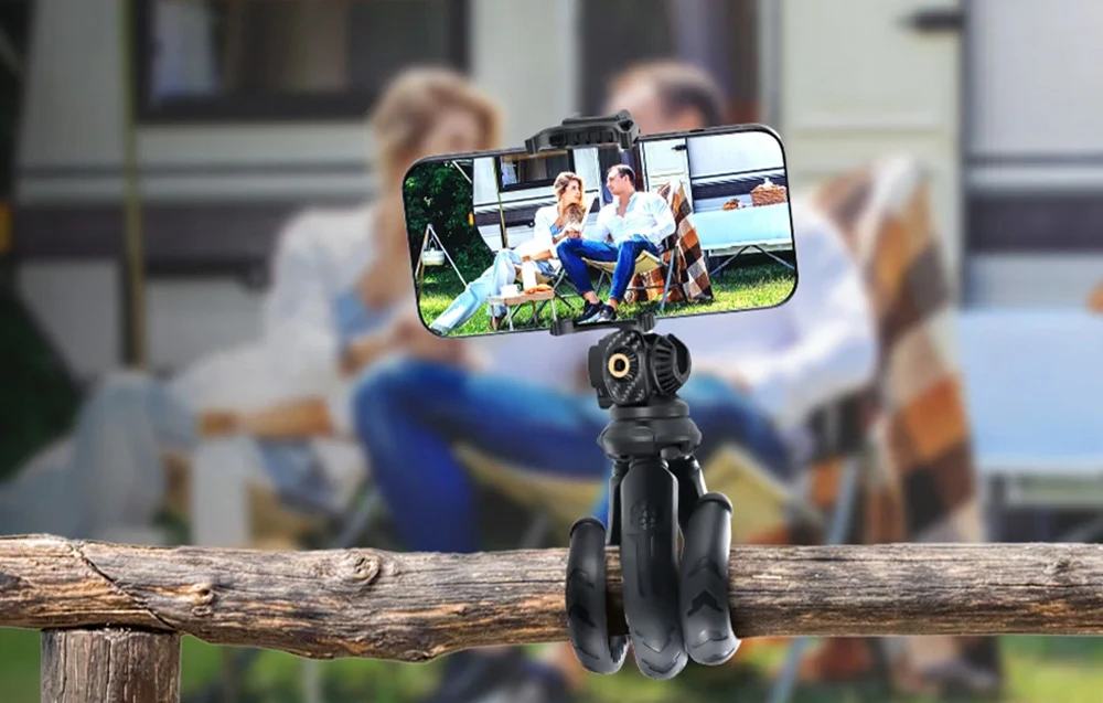 Un trepied flexibil înfășurat în jurul unei balustrade din lemn, susținând un smartphone cu o imagine a unui cuplu într-un camping.