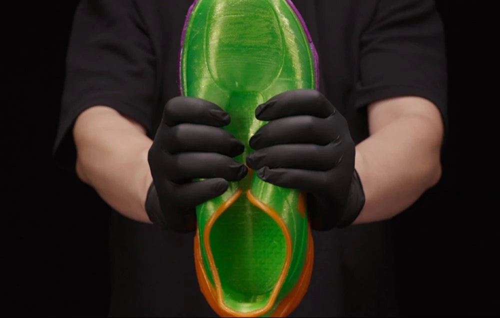 Mâini în mănuși negre strângând un pantof verde, flexibil, imprimat pe o imprimantă 3D