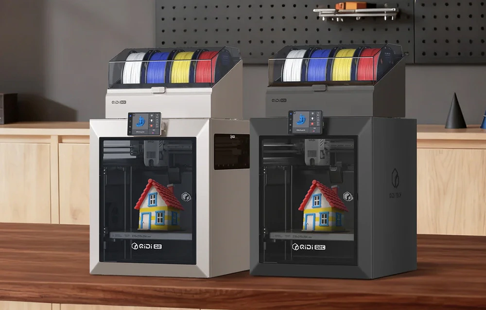 Două imprimante 3D QIDI cu filamente colorate imprimând modele de căsuțe pe o masă de lemn