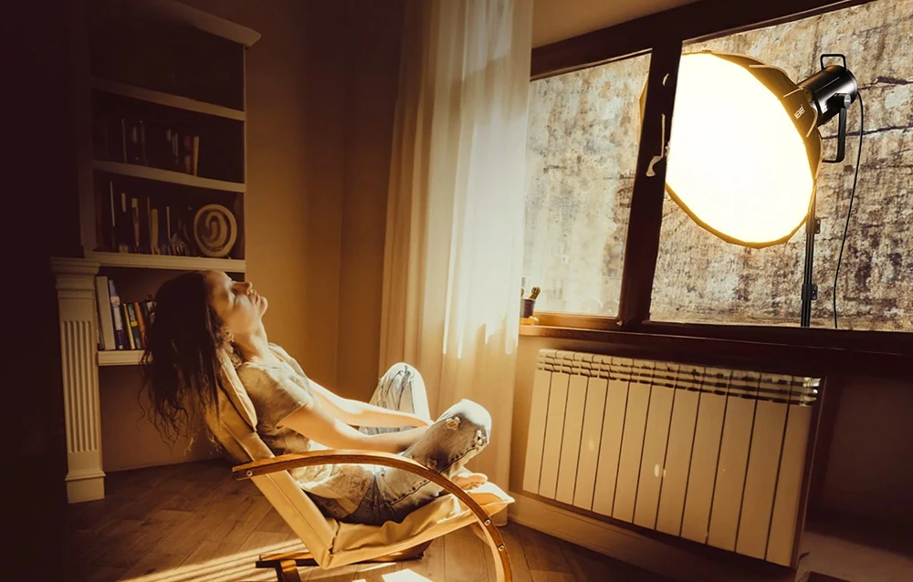 O femeie relaxându-se într-un fotoliu într-o cameră iluminată de lumina caldă a unei lămpi amplasate în spatele ferestrei.