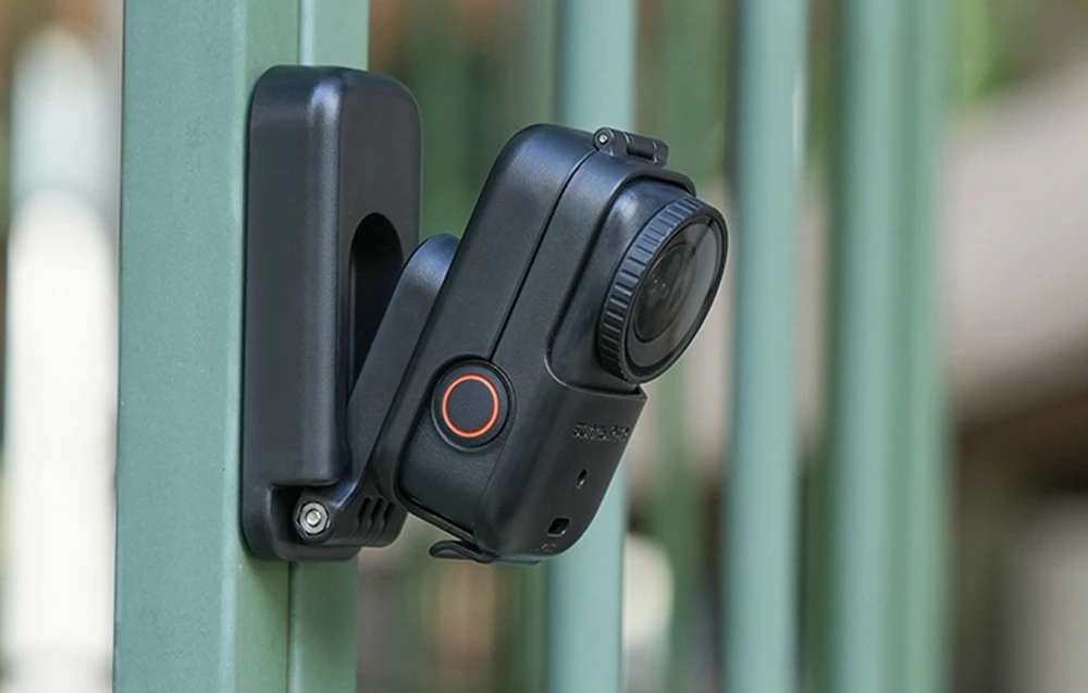 O mini-cameră într-un suport negru atașat vertical la un gard metalic verde.