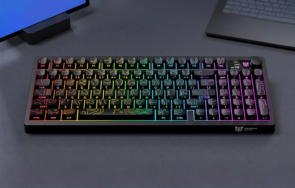 O tastatură cu iluminare colorată așezată pe o masă neagră lângă o tabletă și un laptop