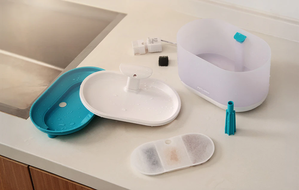 Piesele unui distribuitor de apă pentru pisici așezate pe blatul de bucătărie, inclusiv un rezervor de apă, un capac din plastic, un filtru și accesorii de curățare.